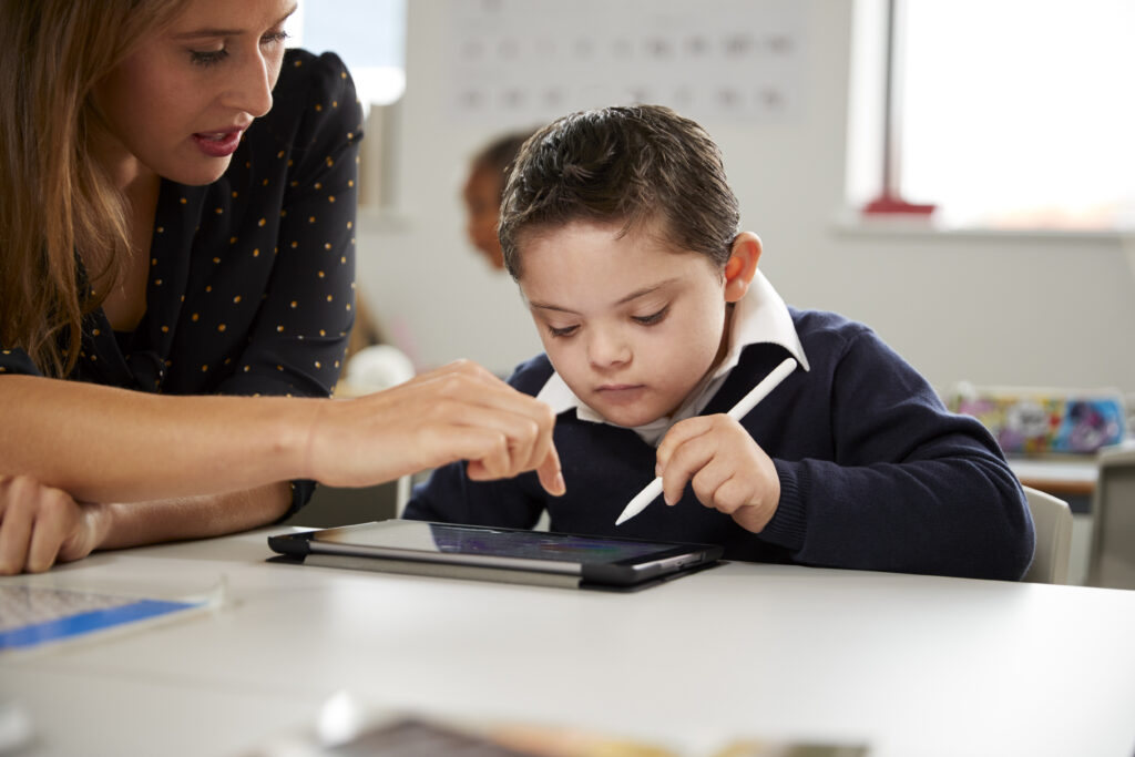 Female teacher working with a boy sitting at desk using a tablet computer in a classroom.
